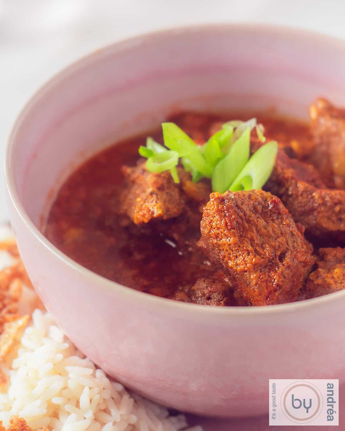 Een hoogtefoto van een roze kom gevuld met curry van rundvlees en kokosmelk. Wat rijst ernaast.