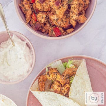 Een vierkante foto genomen van bovenaf. Een roze bord met een wrap gevuld met gyros en salade, een schaaltje met saus en een schaal met meer gyros op een wit marmeren ondergrond.