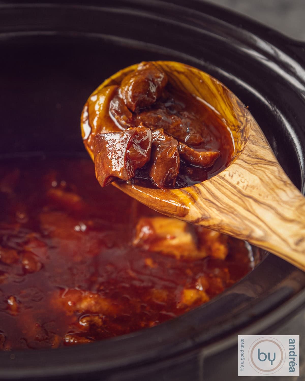 Een slowcooker met een houten lepel waarop blokjes kip liggen. Ajam pangang zit in de slowcooker