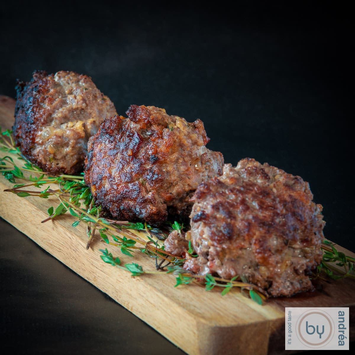 Een vierkante foto met 3 gehaktballen met spek en tijm op een houten plank.