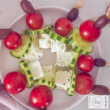 Een vierkante foto met een wit bord met griekse salade spiesjes, komkommer. tomaten, feta en olijven