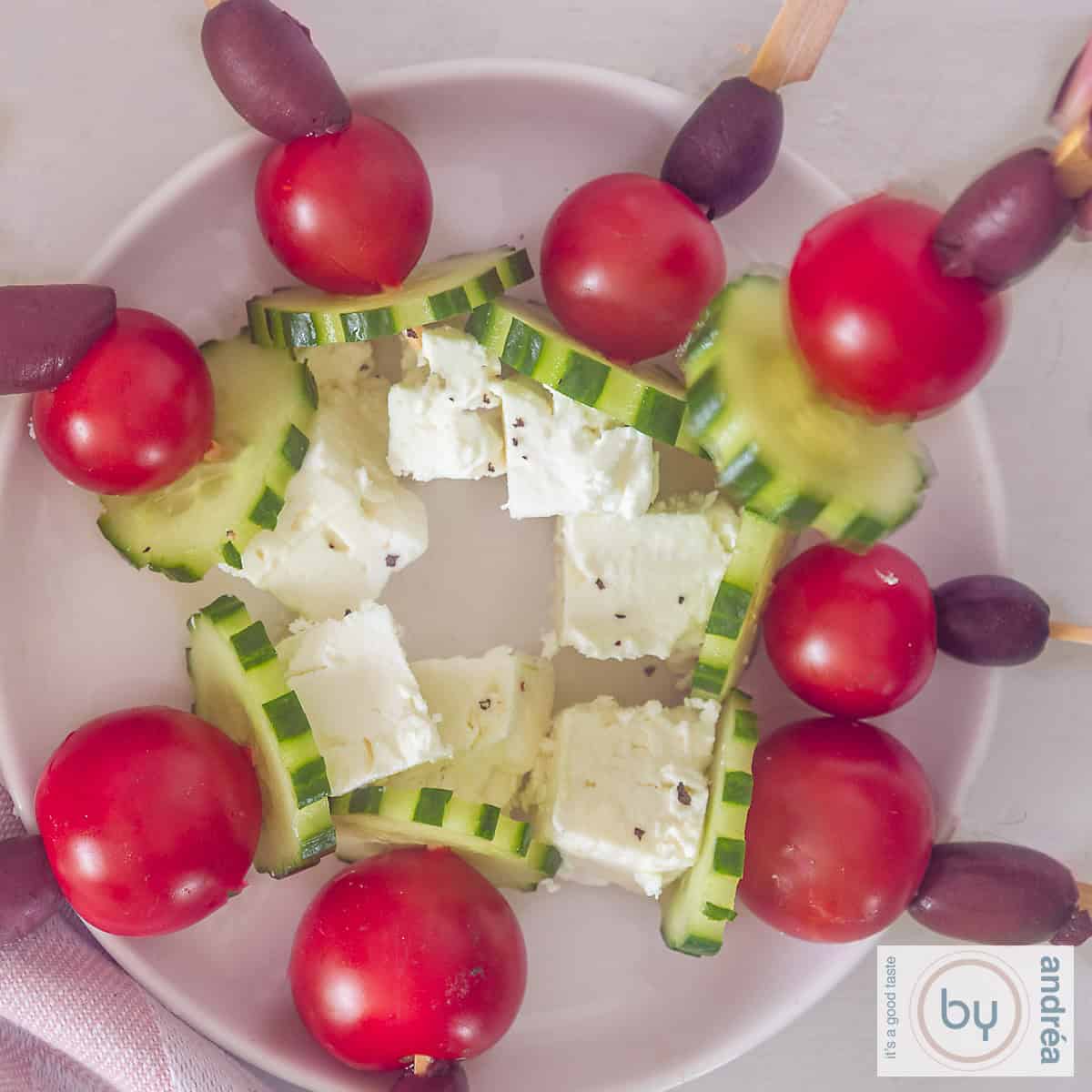 Een vierkante foto met een wit bord met griekse salade spiesjes, komkommer. tomaten, feta en olijven