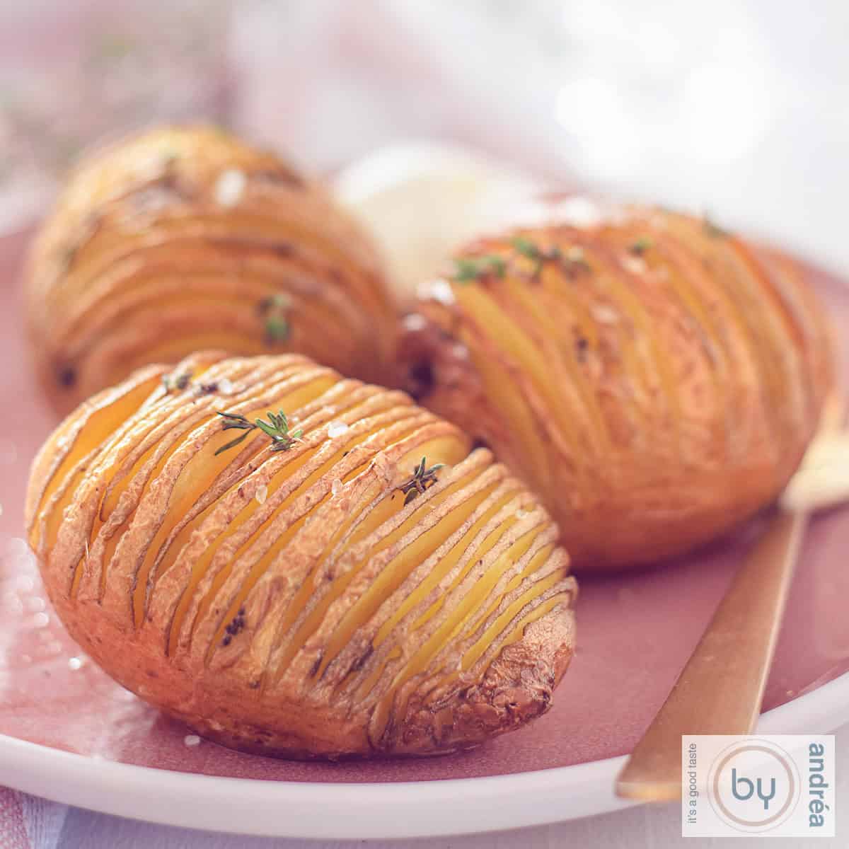 Een vierkante foto met Hasselback potatoes op een roze bord.