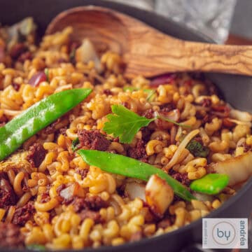 Een pan gevuld met macaroni met een Indische twist, gehakt en groenten. Een houten lepel ligt in de pan.