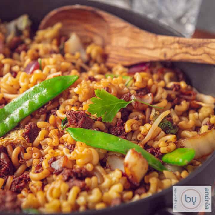 Een pan gevuld met macaroni met een Indische twist, gehakt en groenten. Een houten lepel ligt in de pan.