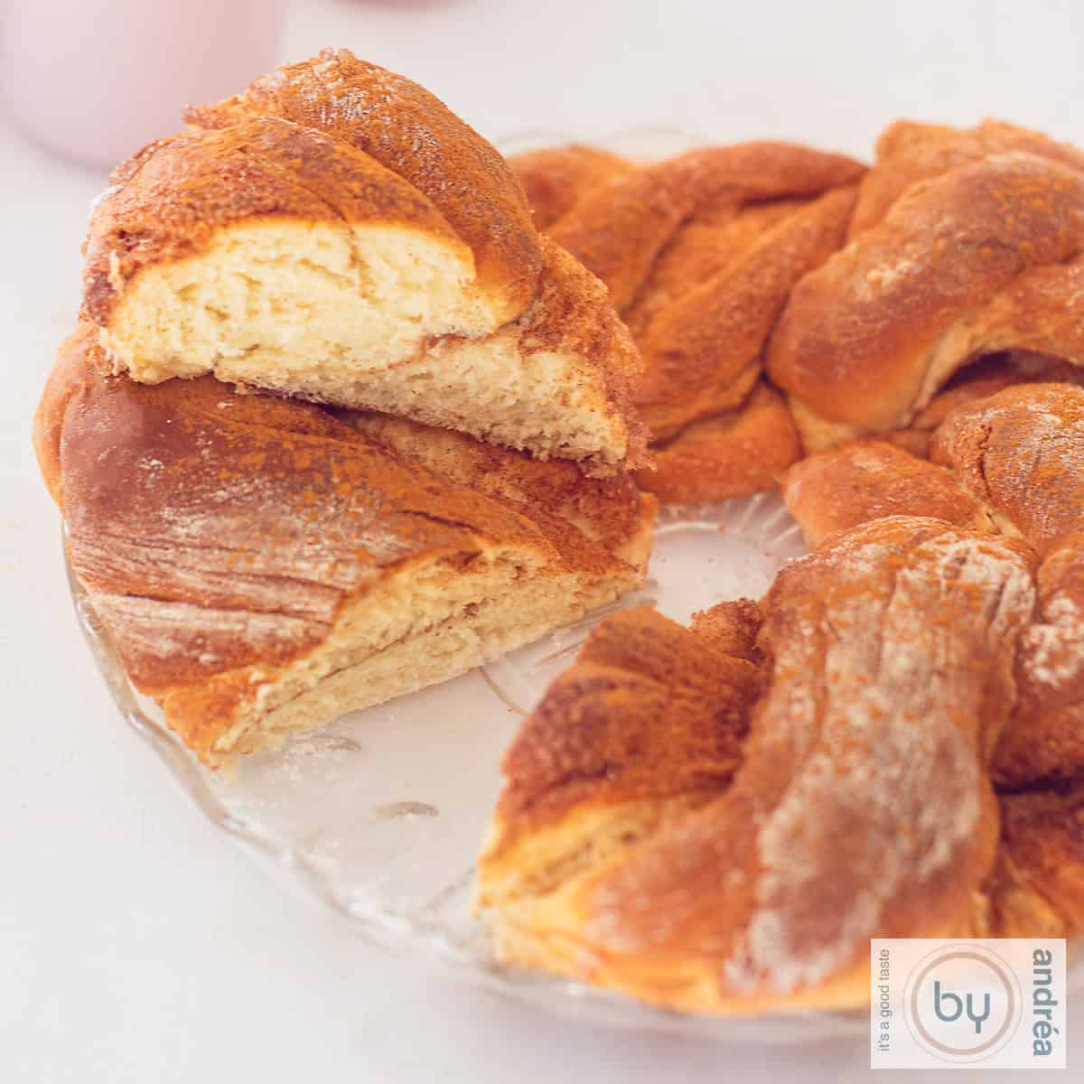 Een vierkante foto met een kaneel brood krans waar een punt is uitgesneden.