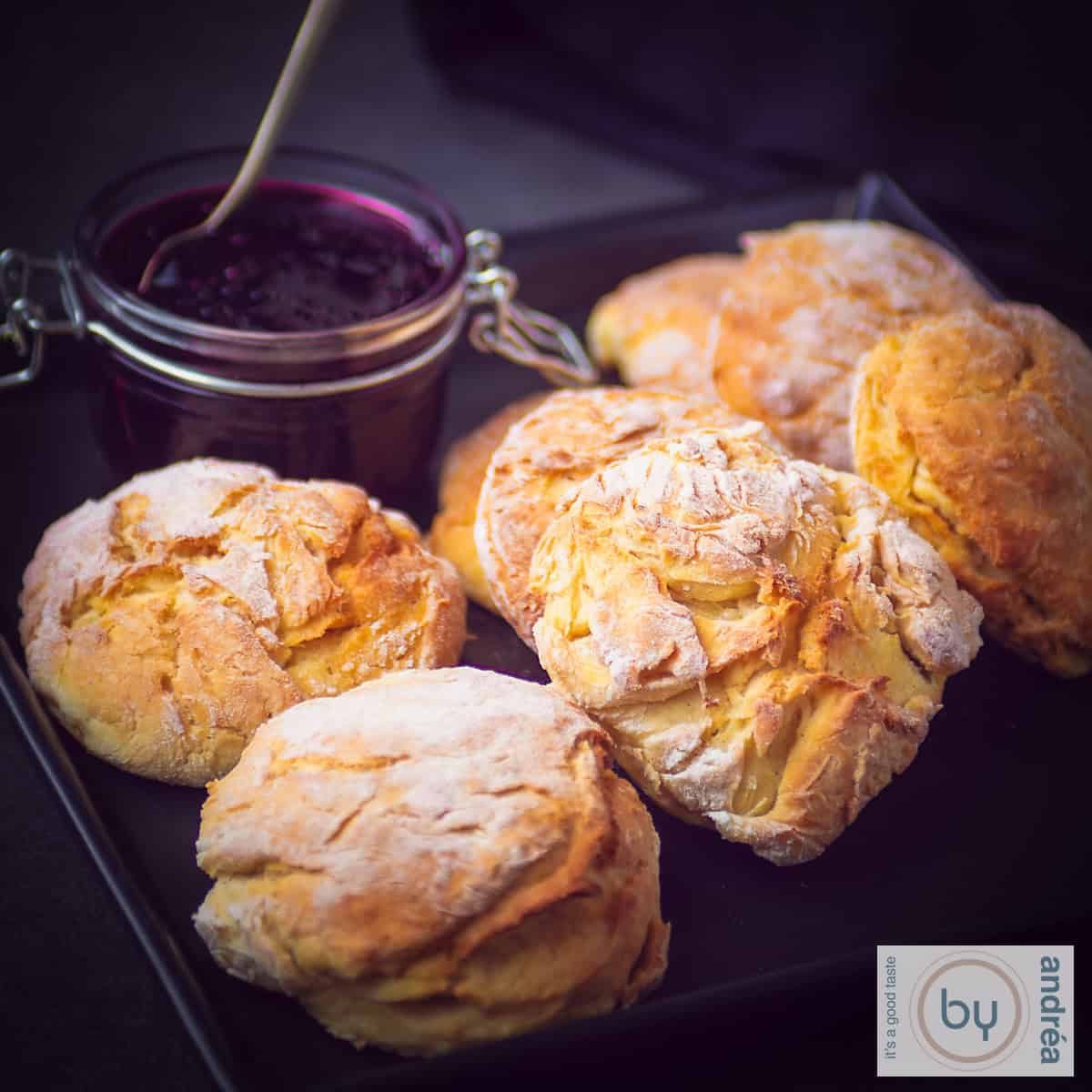Een vierkante foto met 6 scones en braam champagne confituur in een potje