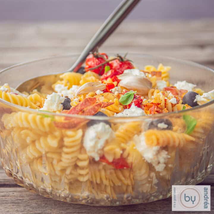Een vierkante foto met een glazen schaal op een houten achtergrond gevuld met pastasalade, tomaatjes, bacon en feta