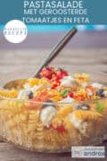 Een deel van een glazen schaal met olijven, feta, geroosterde tomaten, pasta, bacon en basilicum. Een tekst bovenin: pastasalade met geroosterde tomaatjes en feta