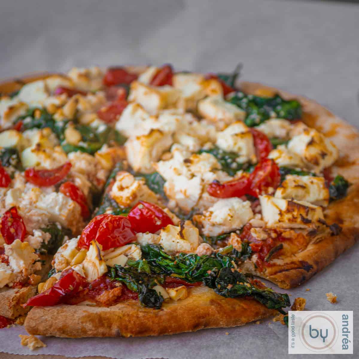 Een vierkante foto met een deel van een pizza belegt met spinazie, feta en tomaat