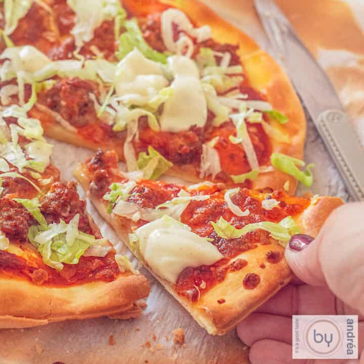 Een vierkante foto met een deel van een pizza. Een hand pakt een punt weg.