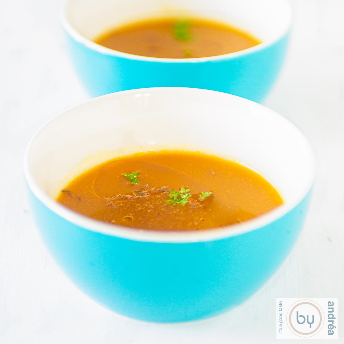 Een witte ondergrond met twee soepkommen achter elkaar gevuld met pompoensoep met saffraan