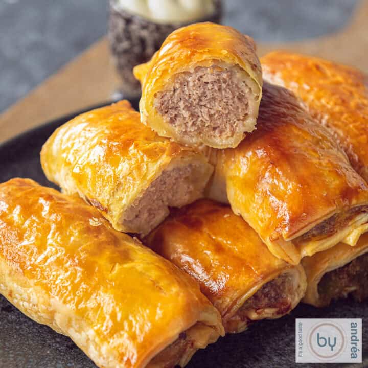 Een vierkante foto met een grijze schaal met een stapel bladerdeeg saucijzenbroodjes. Een broodje gehalveerd