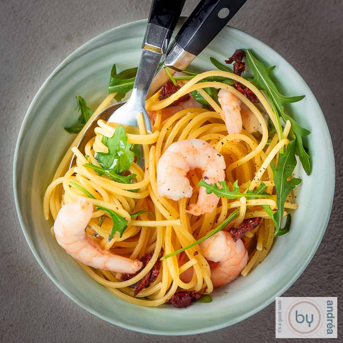 Een vierkante foto met een schaal gevuld met spaghetti, garnalen en rucola