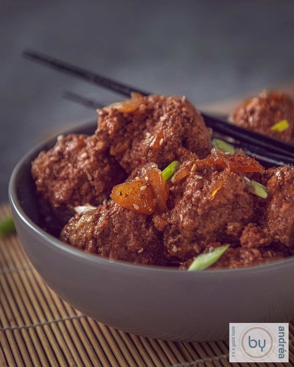 Een grijze schaal met gehaktballetjes in ketjapsaus met twee chopsticks