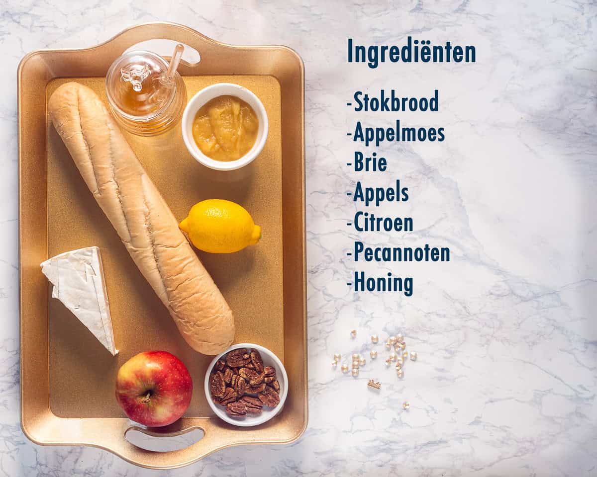 Een gouden dienblad met ingrediënten voor bruschette met brie, walnoot, honing en appel op een marmeren ondergrond met tekstbeschrijving.