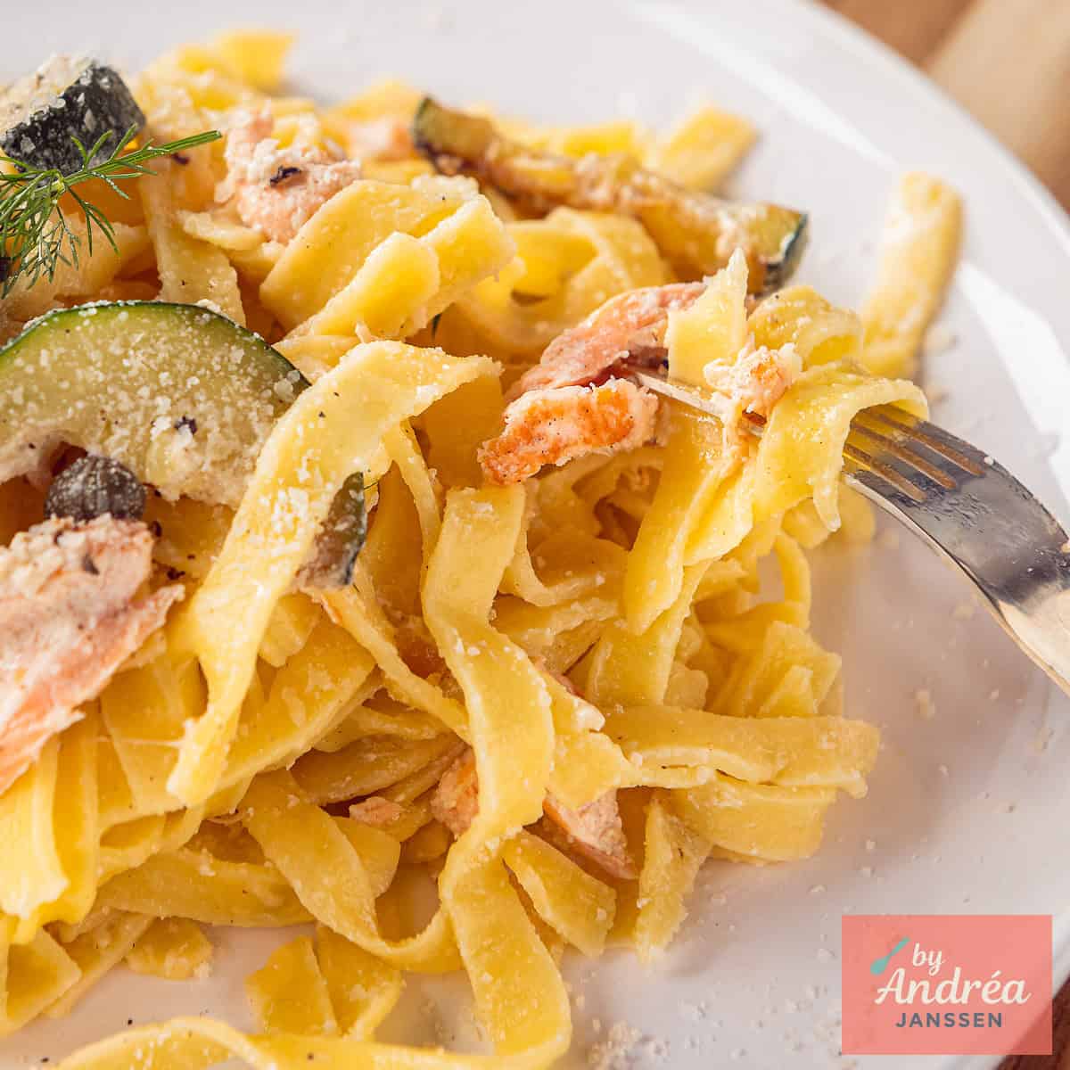 Een vierkante foto met een deel van een wit bord met pasta, courgette, zalm, kappertjes en romige saus.