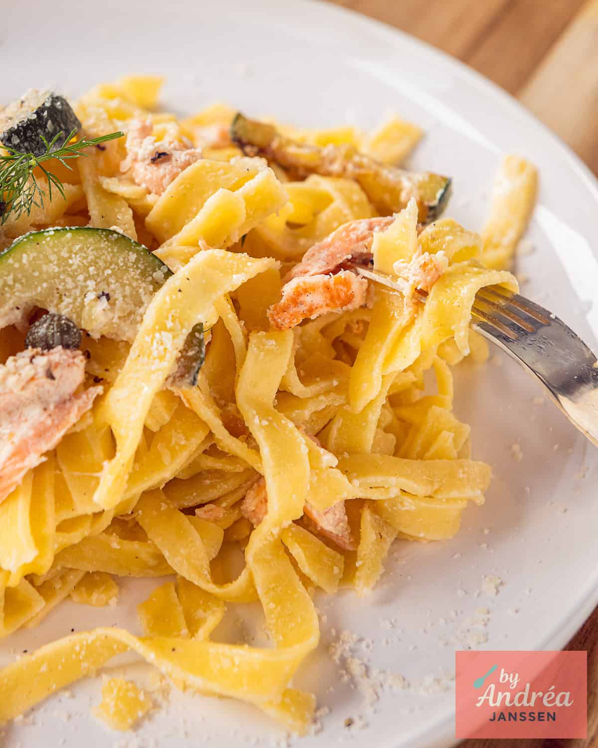 Een deel van een wit bord met pasta, zalm courgette, kappertjes en parmezaanse kaas.