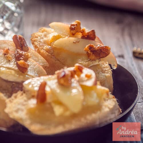 Een zwart schaaltje met drie plakjes stokbrood met honing, appel, Brie en walnoot