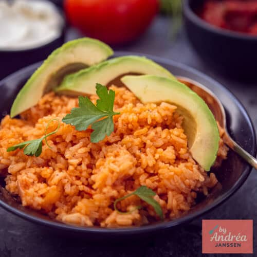 Een vierkante foto met een zwarte schaal met rode Mexicaanse rijst met avocado en peterselie