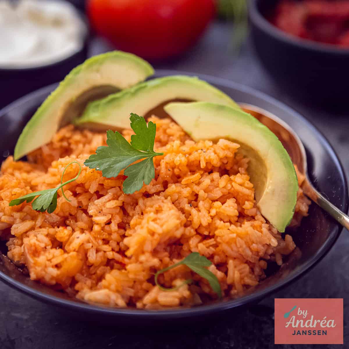 Een vierkante foto met een zwarte schaal met rode Mexicaanse rijst met avocado en peterselie