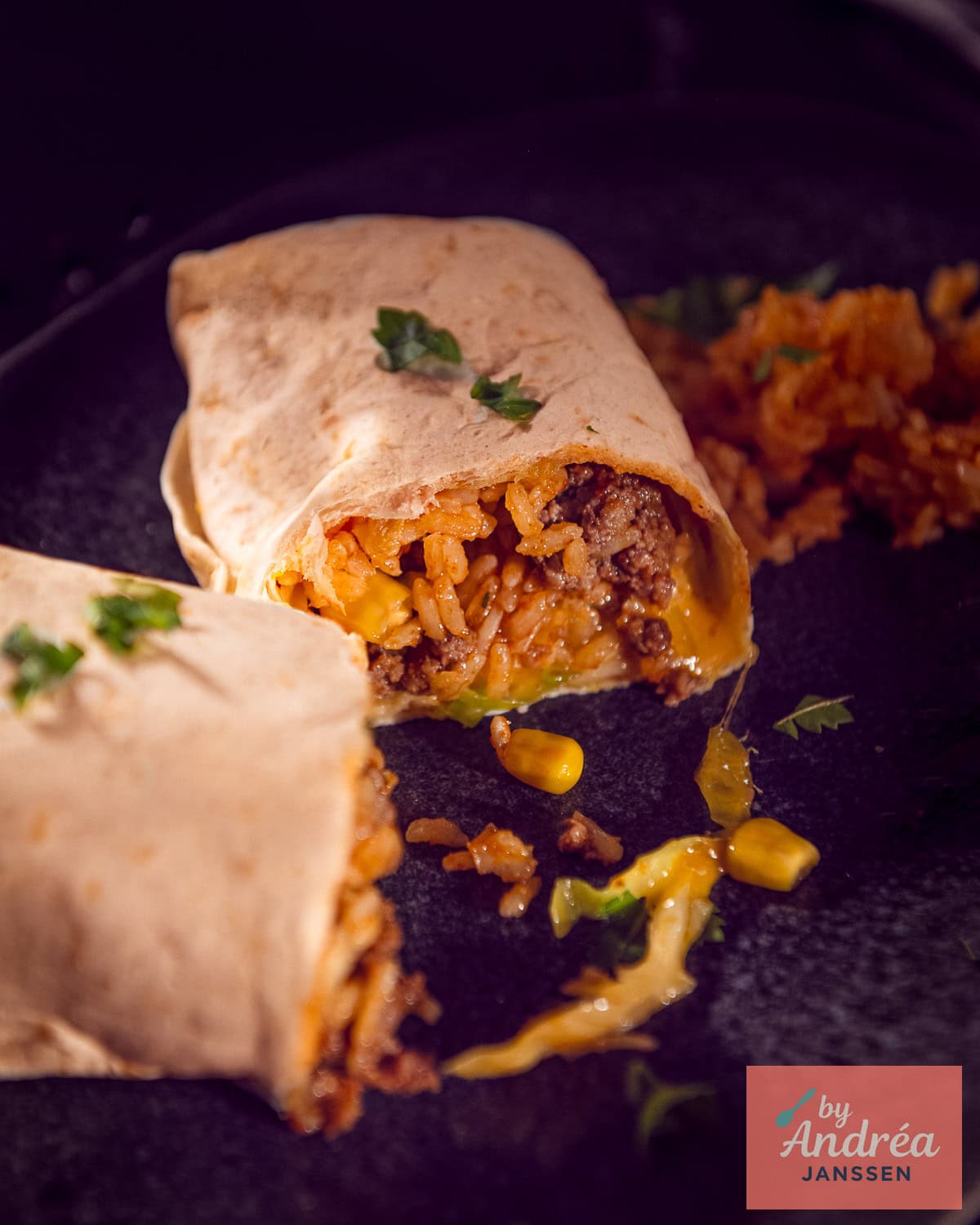 Een grijs bord met een burrito opengesneden zodat je de vulling met rijst, gehakt, mais, kool, tomaat en kaas goed kunt zien.