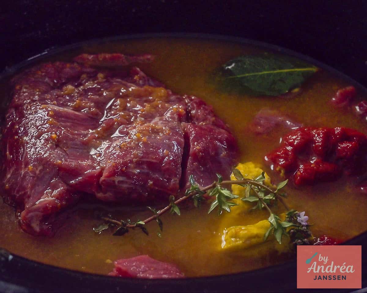 Een slowcooker gevuld met rundvlees, bouillon, mosterd, tomatenpuree, kruiden en andere smaakmakers