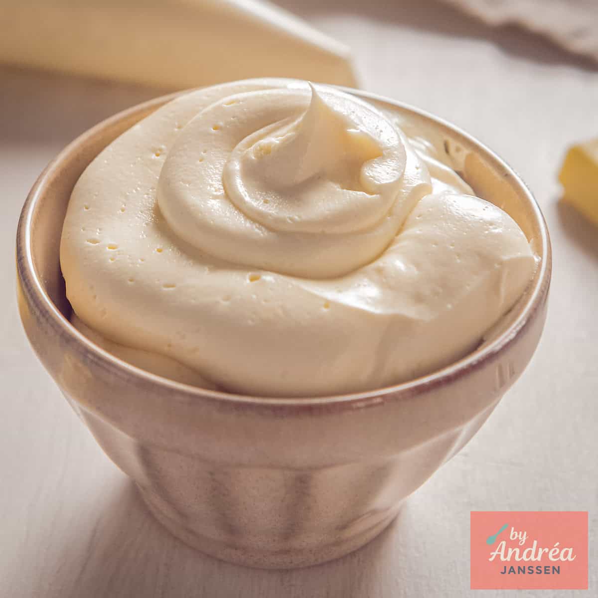 Een vierkante foto met een beige kom met vanille witte chocolade botercrème.