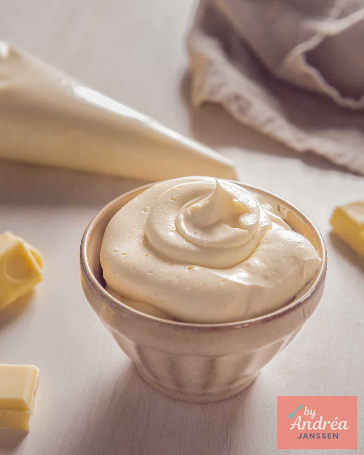 Een beige kom met Romige Witte Chocolade Botercrème. In de achtergrond een spuitzak met meer crème