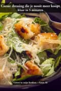 Close-up van caesar salade in een donkere kom met goudbruine krokante croutons, geraspte Parmezaanse kaas en romige dressing over frisgroene romeinse sla, met een vork aan de rand van de kom.