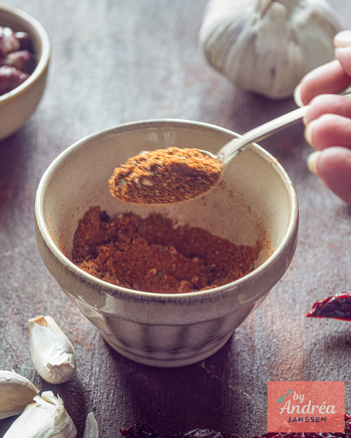 Close-up van een lepel vol chili con carne kruidenmix boven een kom, met knoflook, kidneybonen en gedroogde chilipeper eromheen.