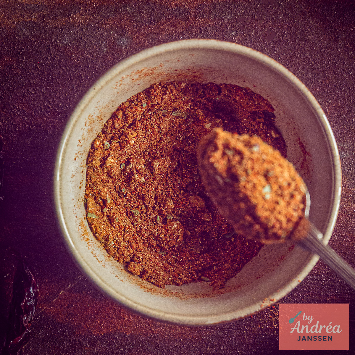 Bovenaanzicht van een kom met roodbruine chili con carne kruidenmix en een lepel vol kruiden.