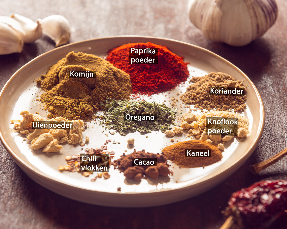 Bord met gelabelde ingrediënten voor chili con carne kruidenmix: komijn, paprikapoeder, koriander, oregano, uienpoeder, knoflookpoeder, chilivlokken, cacao en kaneel.