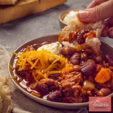 Close-up van chili con carne uit de slowcooker met gehakt, bonen, geraspte cheddar, zure room en brood.