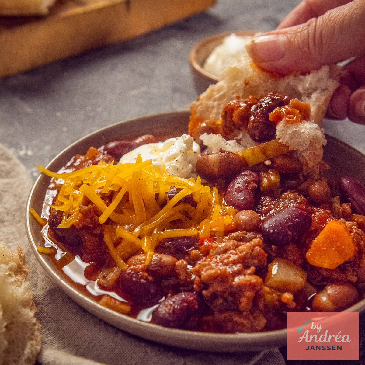 Close-up van chili con carne uit de slowcooker met gehakt, bonen, geraspte cheddar, zure room en brood.