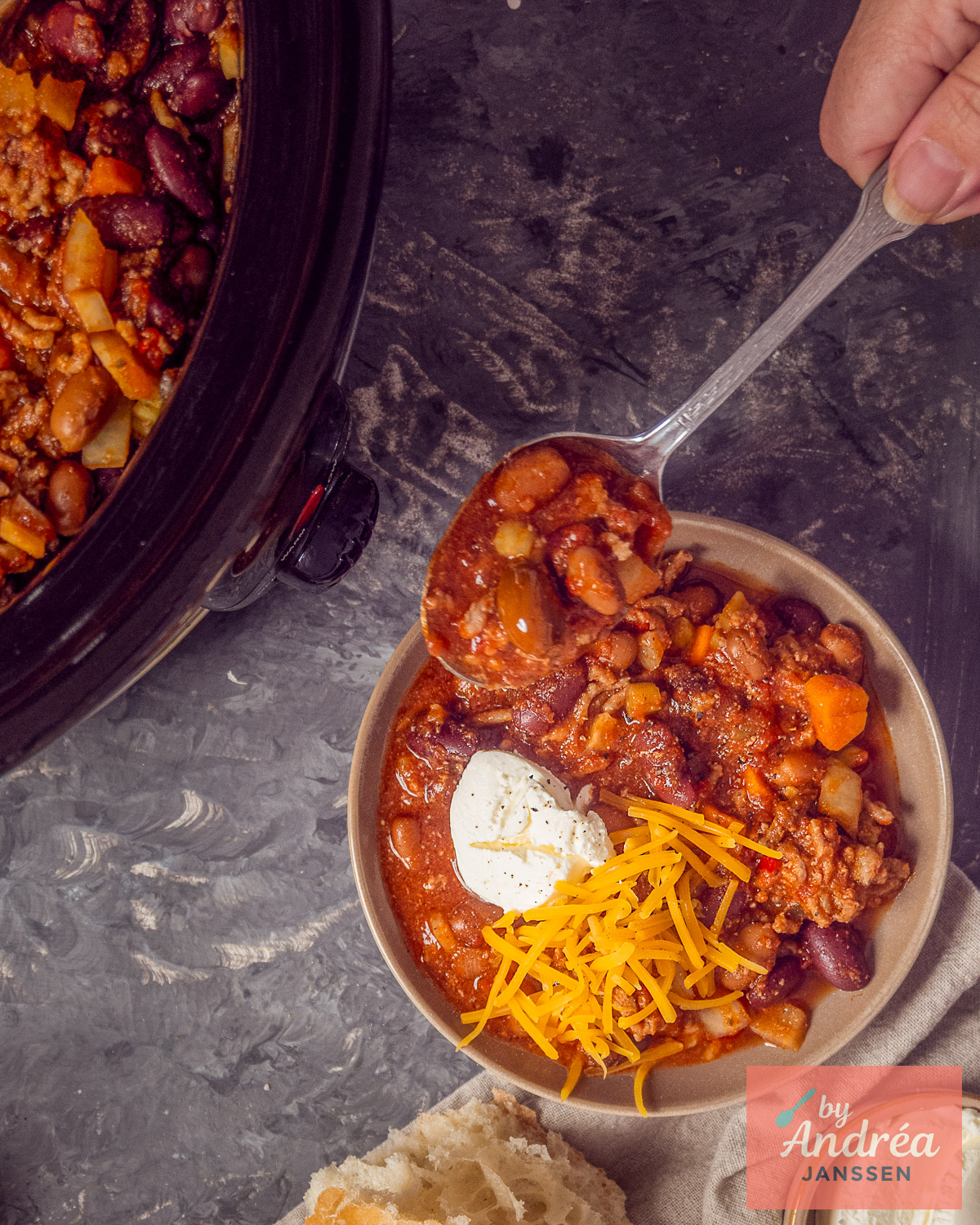 Lepel vol chili con carne met gehakt, kidneybonen, bruine bonen en groenten boven een kom met cheddar en zure room.