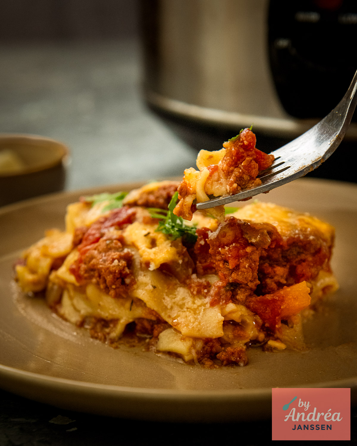 Een vork tilt een hap lasagne met gehaktsaus en kaas op boven een bord, met de slowcooker op de achtergrond.