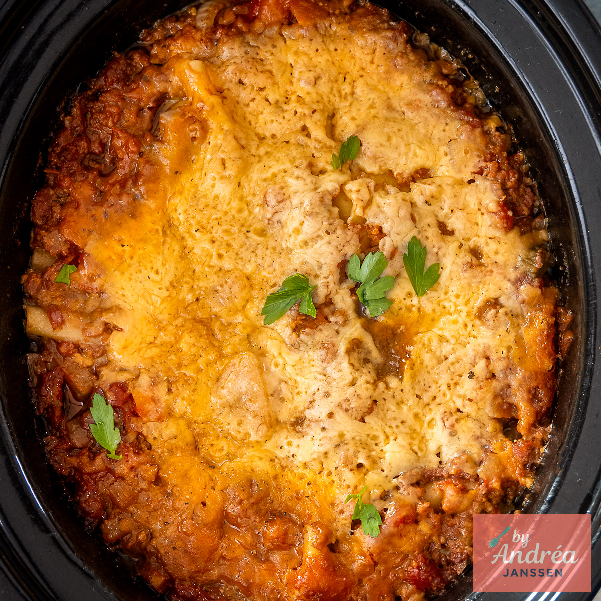Bovenaanzicht van lasagne met een goudgele kaaslaag en peterselie in een zwarte slowcooker.