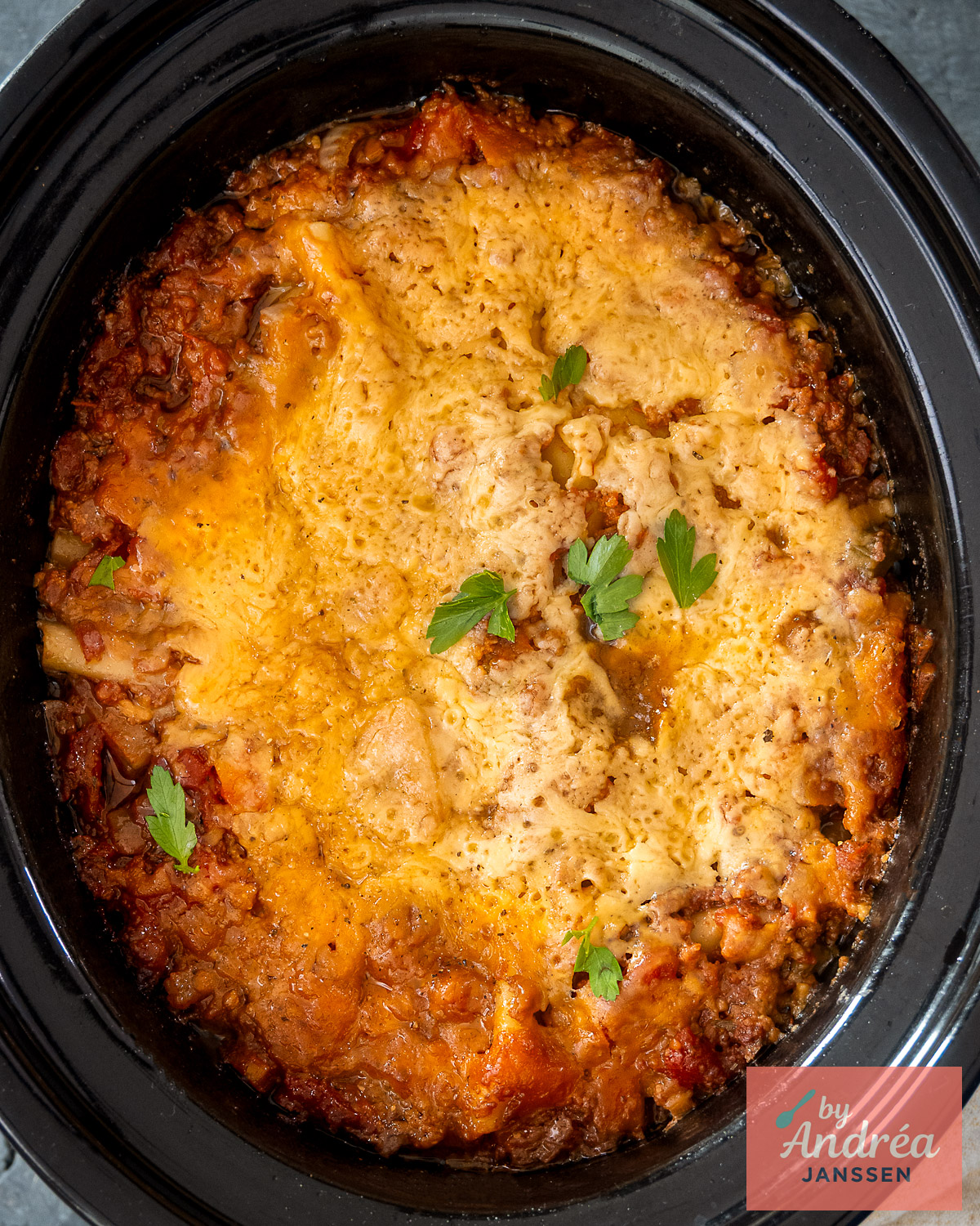 Bovenaanzicht van lasagne met een goudgele kaaslaag en peterselie in een zwarte slowcooker.