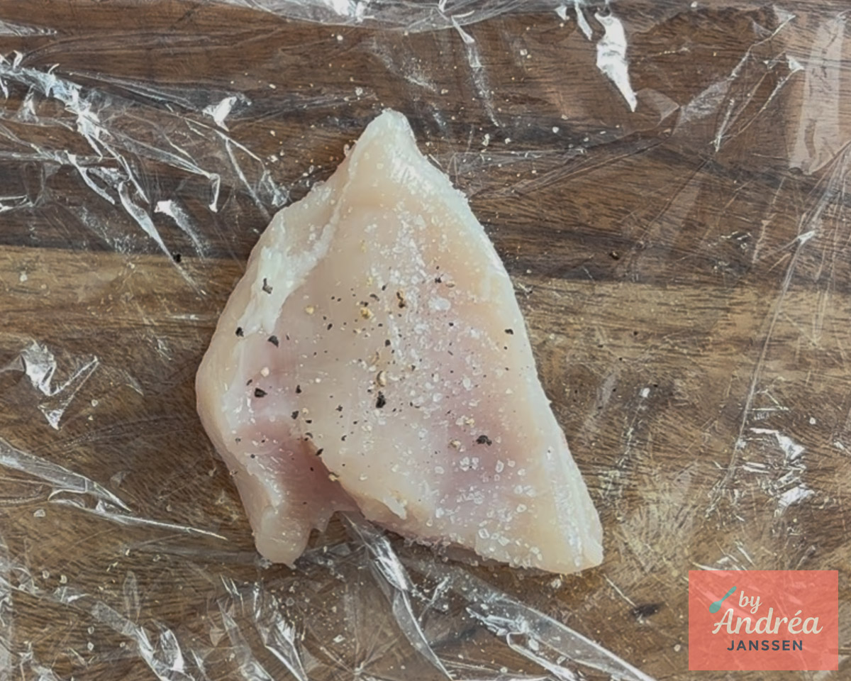 Rauwe kipfilet bestrooid met zout en peper op vershoudfolie op een houten snijplank.