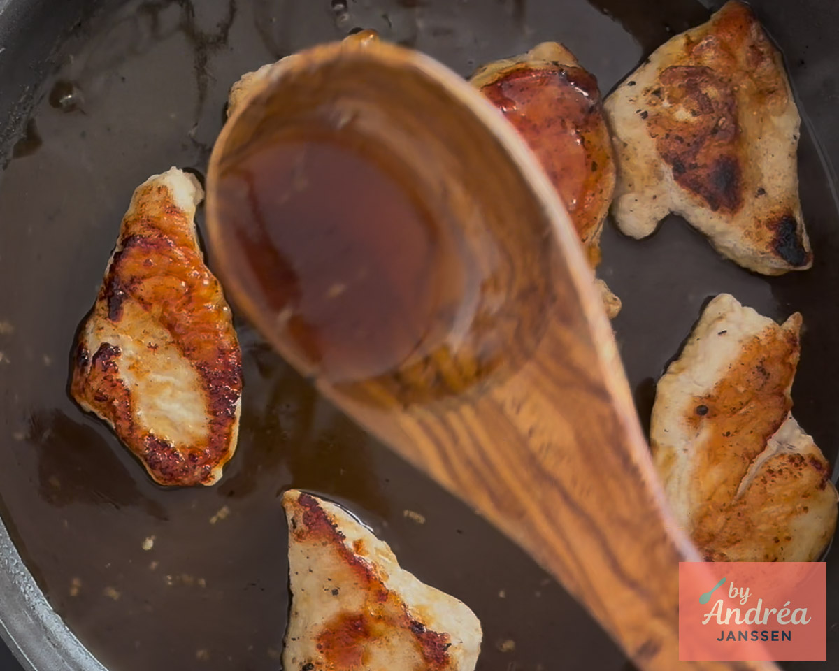 Een houten lepel met honing-citroensaus boven goudbruin gebakken kipfilets in de pan.