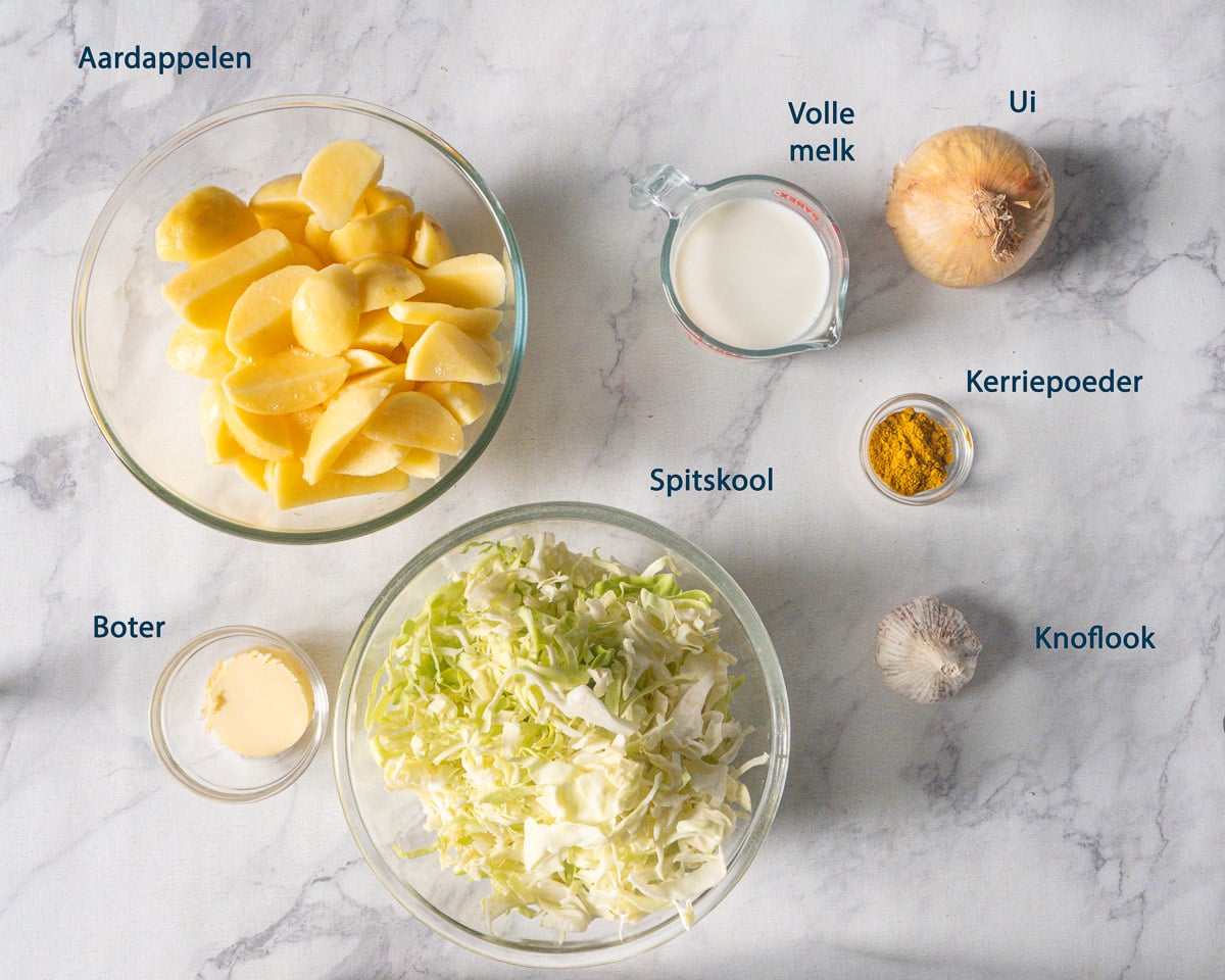 Aardappelen, gesneden spitskool, volle melk, ui, kerriepoeder, boter en knoflook met Nederlandse labels op een lichte ondergrond.