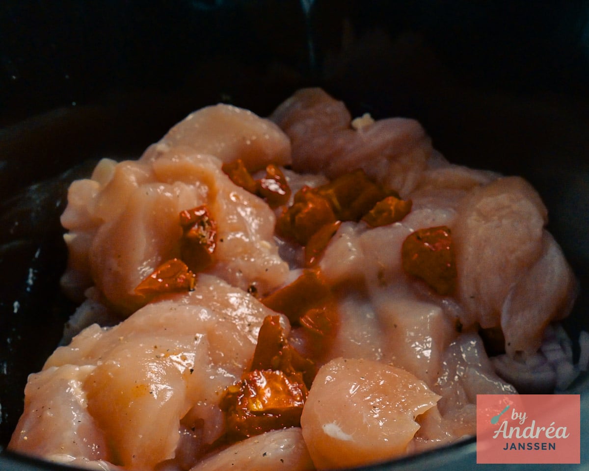 Rauwe kipblokjes met stukjes zongedroogde tomaat op een laag gesnipperde sjalot en knoflook in de slowcooker.
