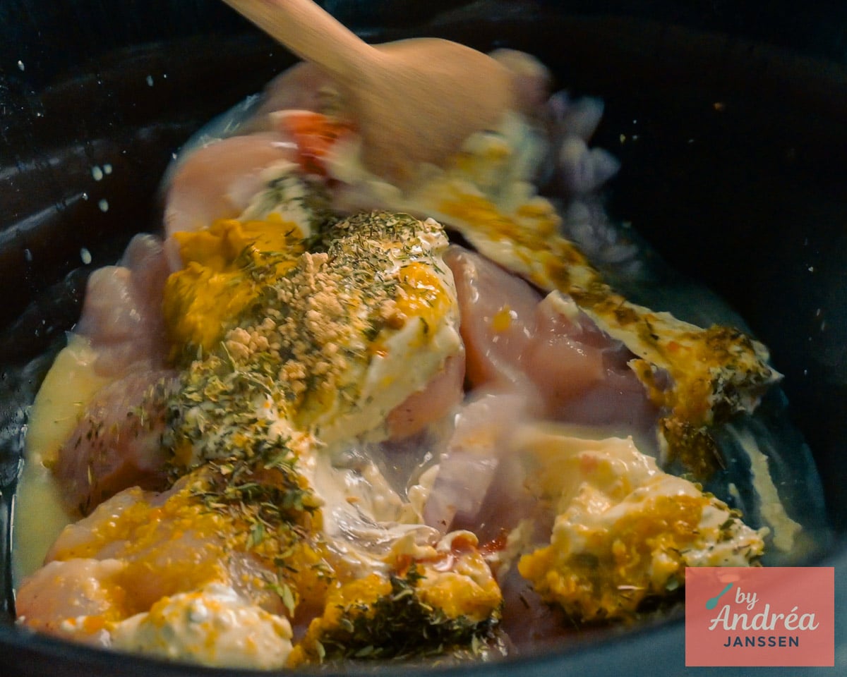 Een houten lepel mengt rauwe kip met Boursin, Dijonmosterd, gedroogde kruiden en vocht in de slowcooker.
