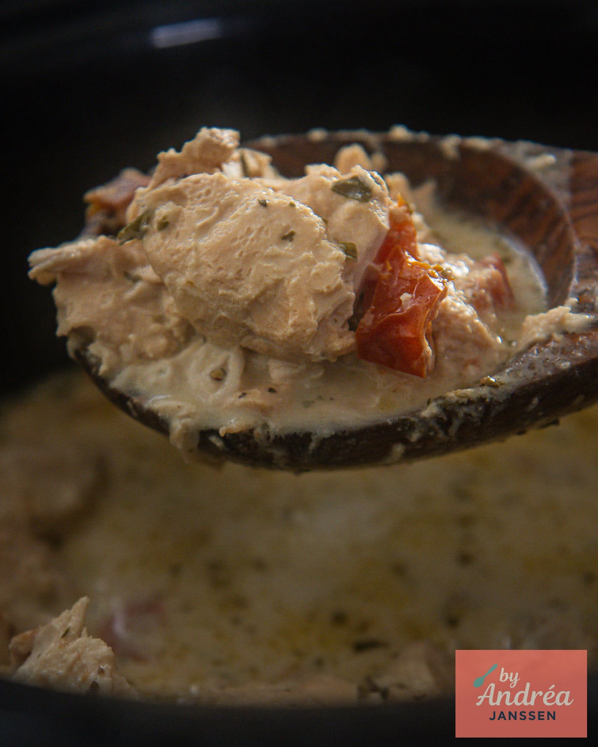 Houten lepel met stukjes kip en zongedroogde tomaat in romige Boursinsaus boven de slowcooker.